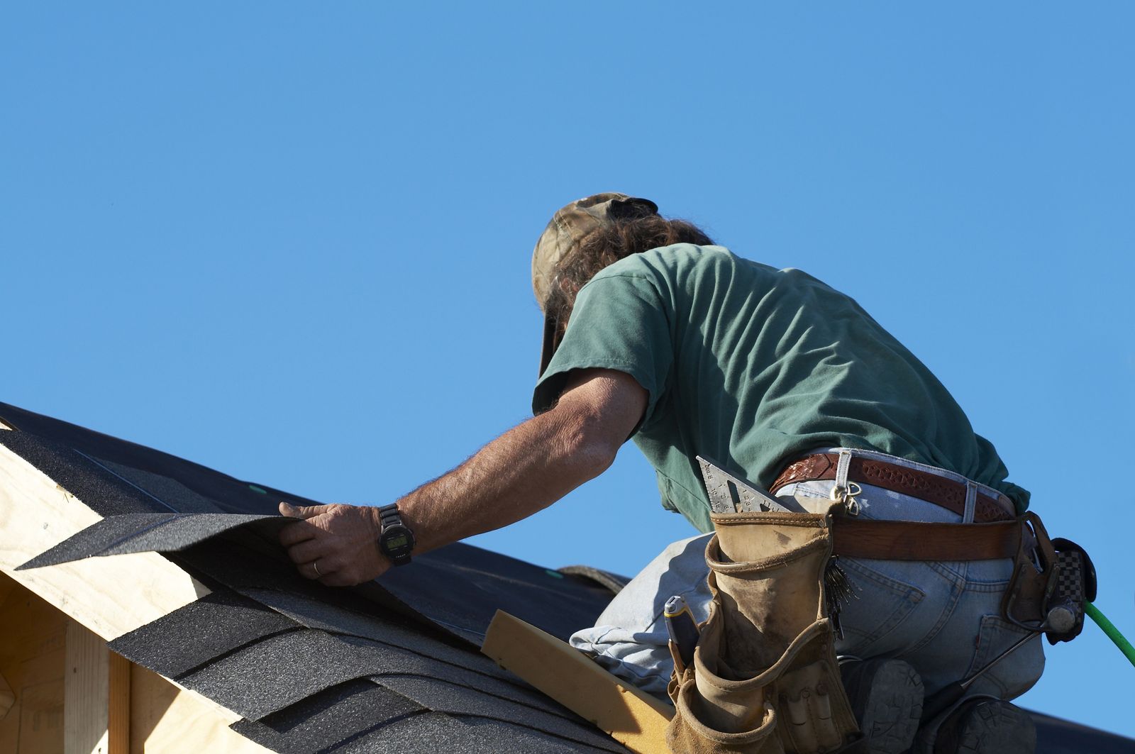 Roofer kneeling on a shingle roof under a clear sky, cutting a shingle to fit